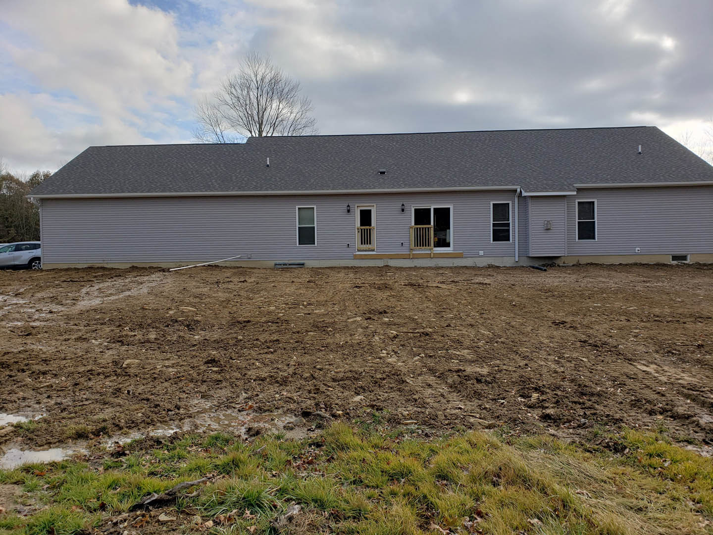 Grey cottage-style home with wooden porch railing, dirt field and sparse grass in foreground, leafless tree beside house, cloudy sky overhead, parked car with damaged side near