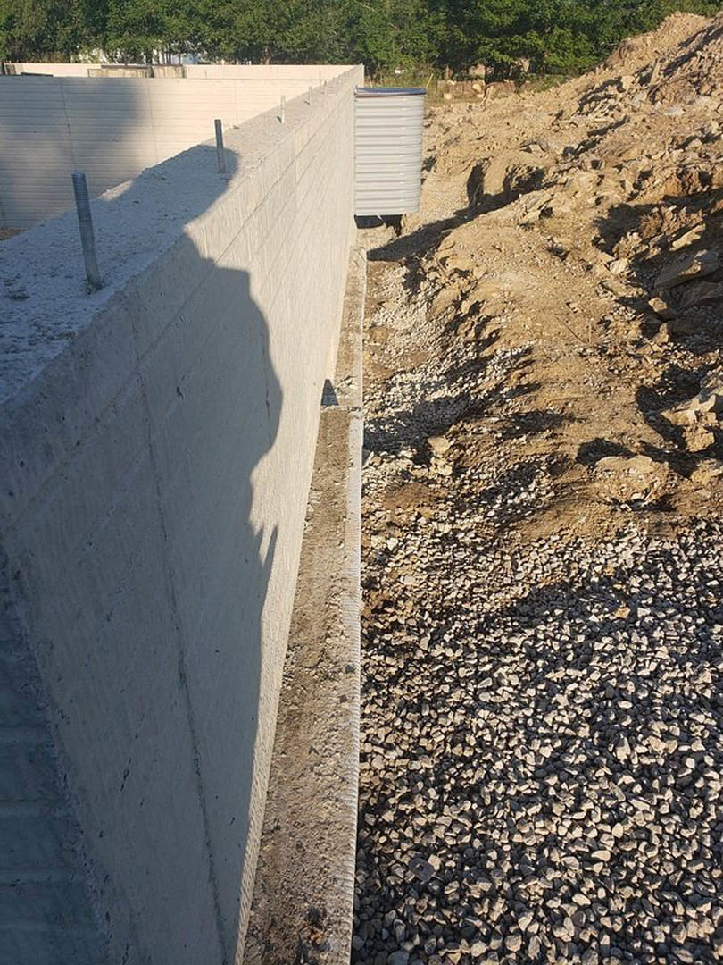 Concrete wall with exposed metal rods, shadow of a person and pole cast on the surface, small rocks and grass field in the foreground, white container nearby.