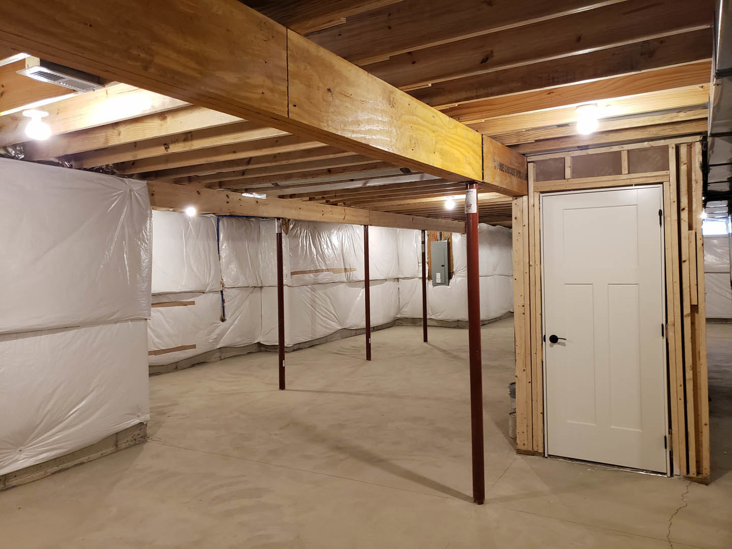 Room under construction with white plastic sheeting covering walls and floor, exposed wooden ceiling beam, white door with black handle, stack of plastic material, and brown