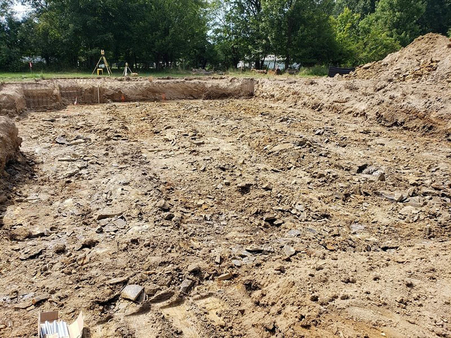 Dirt ground with a large excavation hole, surrounded by scattered rocks and a tripod, bordered by trees under a clear sky