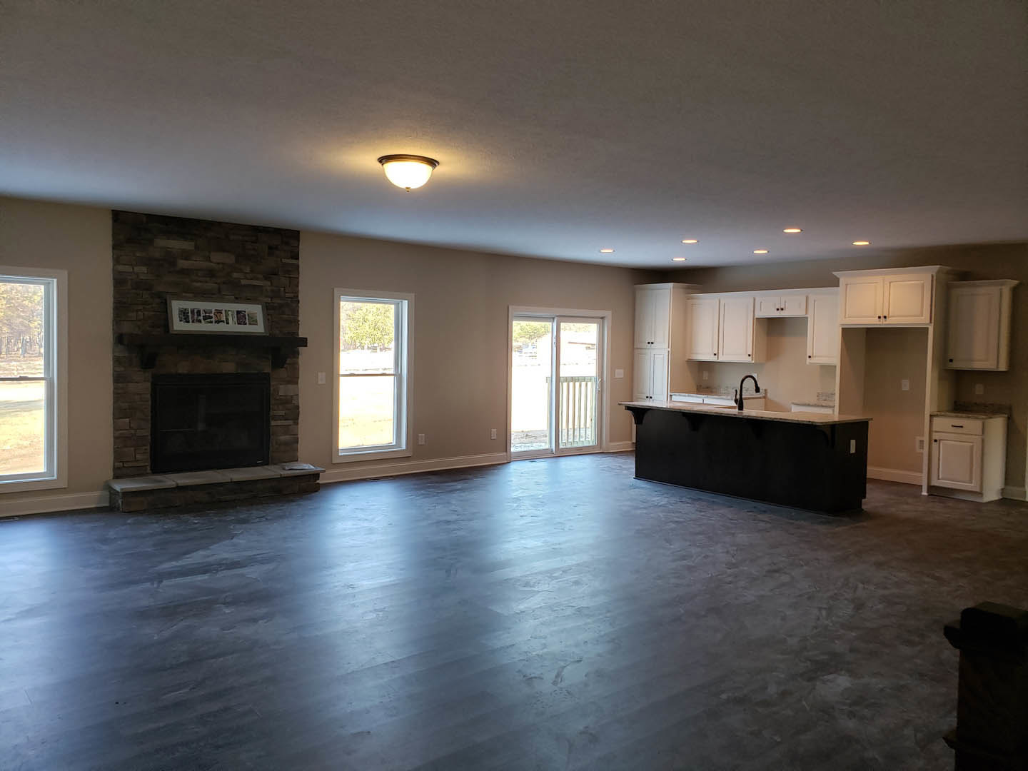 Spacious living room featuring dark wood flooring, stone fireplace, black countertop with faucet, sliding glass door with white railing, large window overlooking trees, and framed