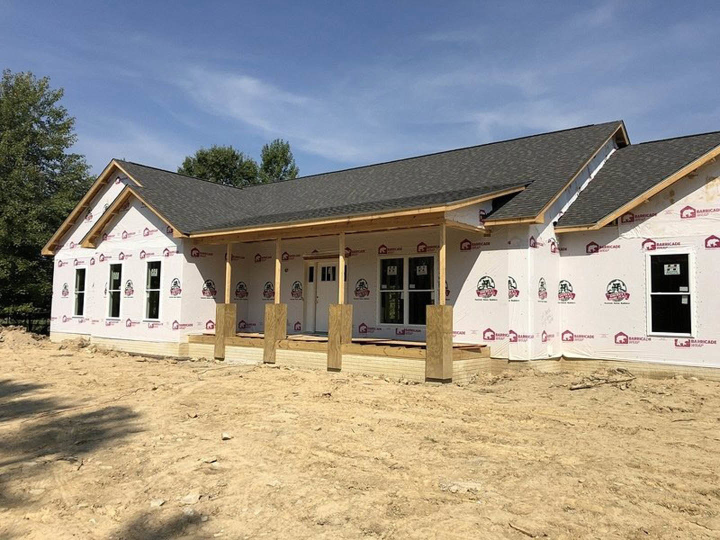 Partially built house with exposed wooden framing, windows covered with white paper signs, unfinished roof, and dirt ground surrounded by scattered wooden pillars.
