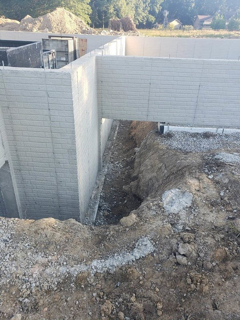 Excavated hole in soil beside a white brick exterior wall, with construction materials visible around foundation area