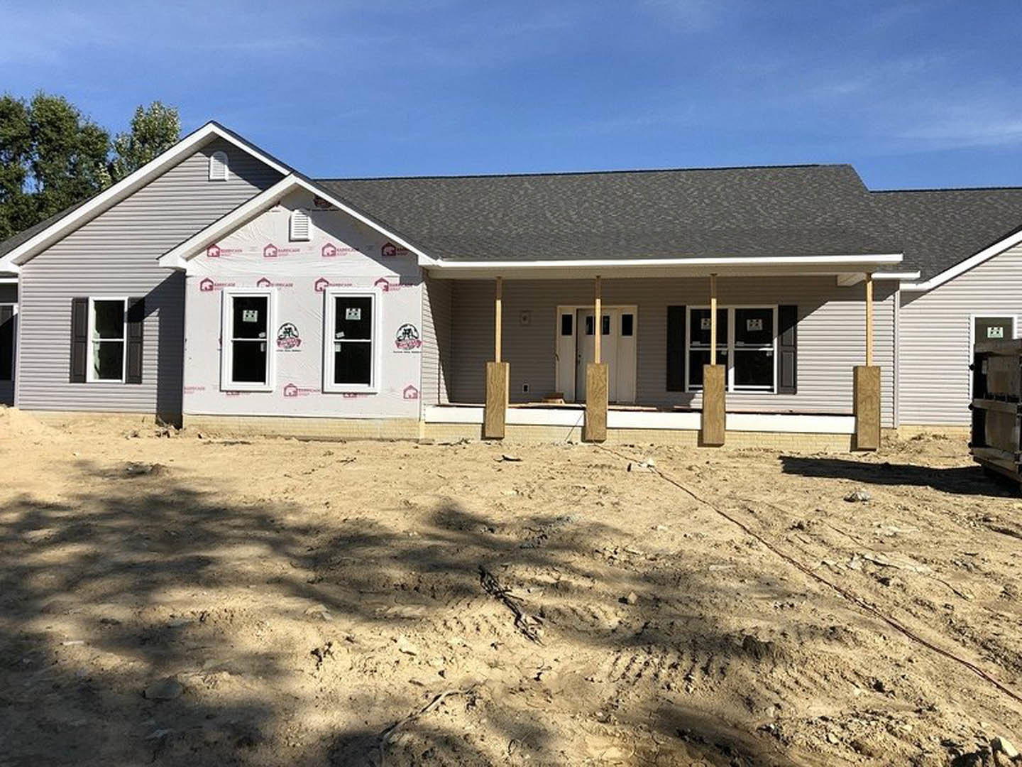 Modern home exterior with prominent wooden posts and beams, white-framed windows with stickers, unfinished dirt ground, and tree shadow across the front.