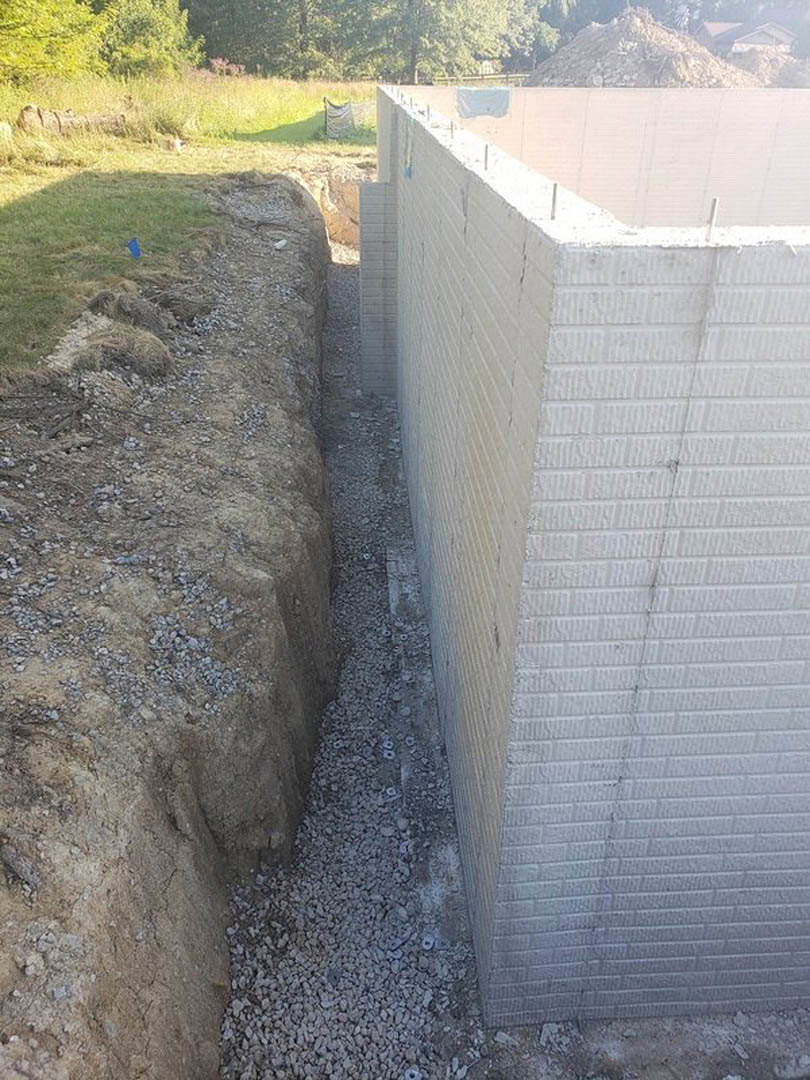 White brick exterior wall with a circular hole in the ground, surrounded by concrete and dirt, with trees visible in the background.