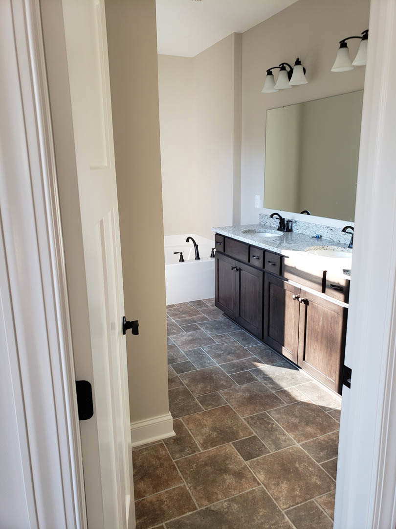 Bathroom with two undermount sinks set in a stone countertop, freestanding bathtub, large wall mirror, three-light fixture above sinks, and light-colored tile flooring