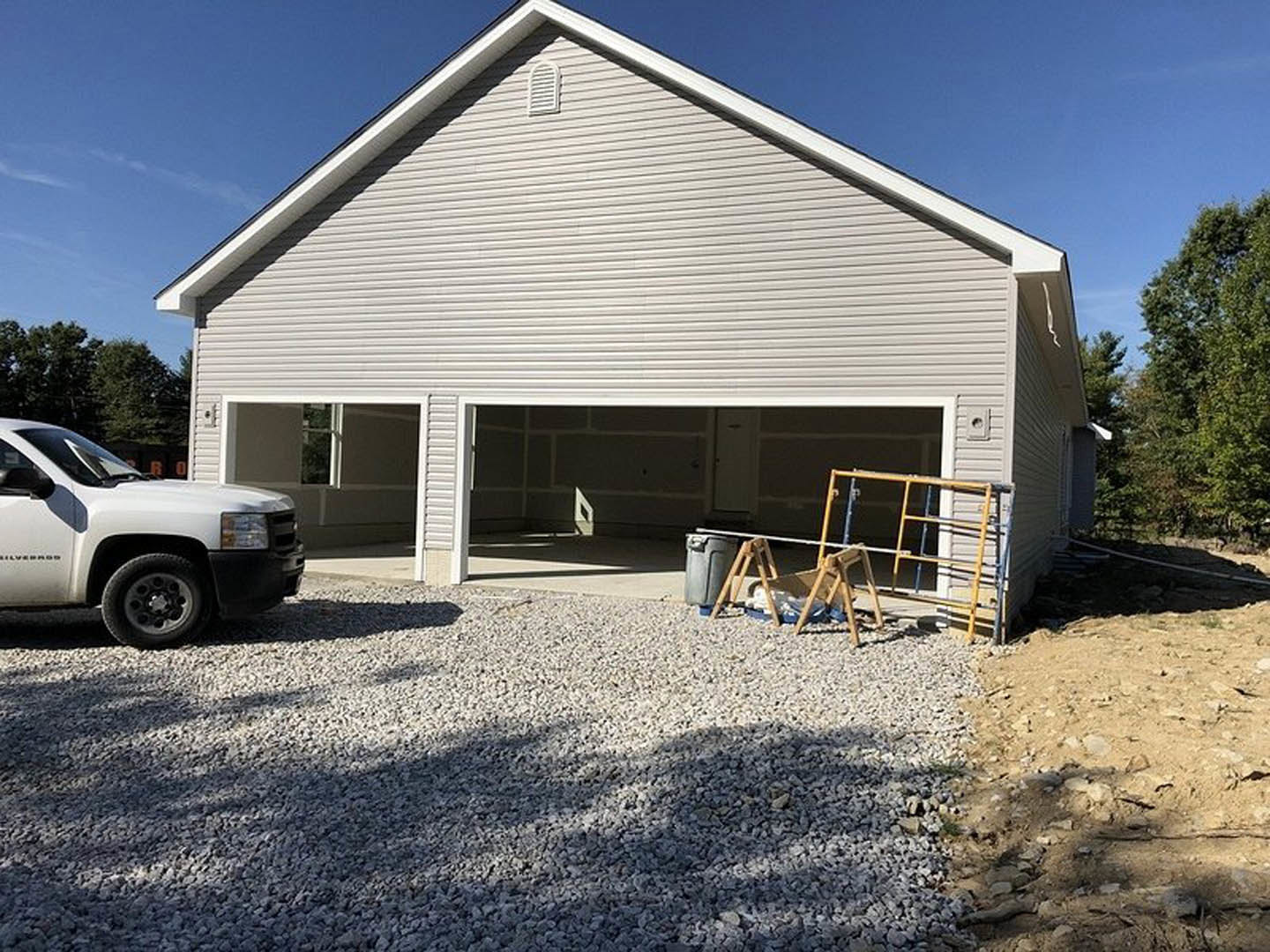 Garage with white truck parked on gravel, yellow scaffolding beside building, garage door partially visible, child sitting in sled, trees in background