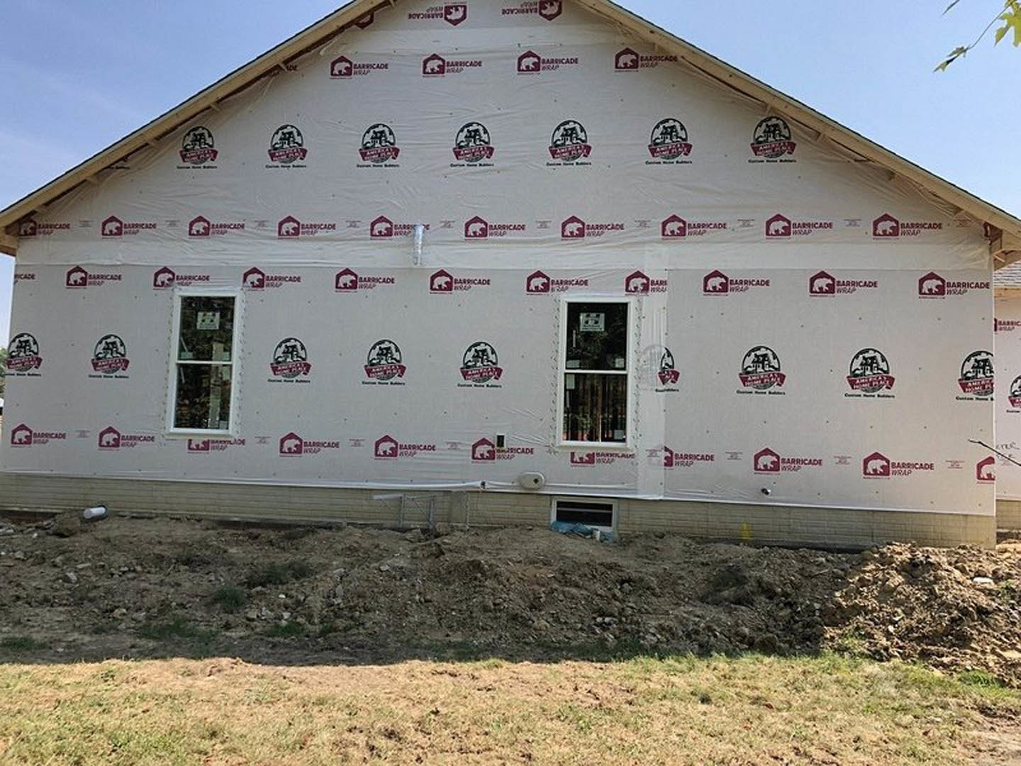 Two-story house wrapped in white plastic sheeting, white-framed window with sign, grassy yard with person standing, pile of dirt near foundation, overcast sky