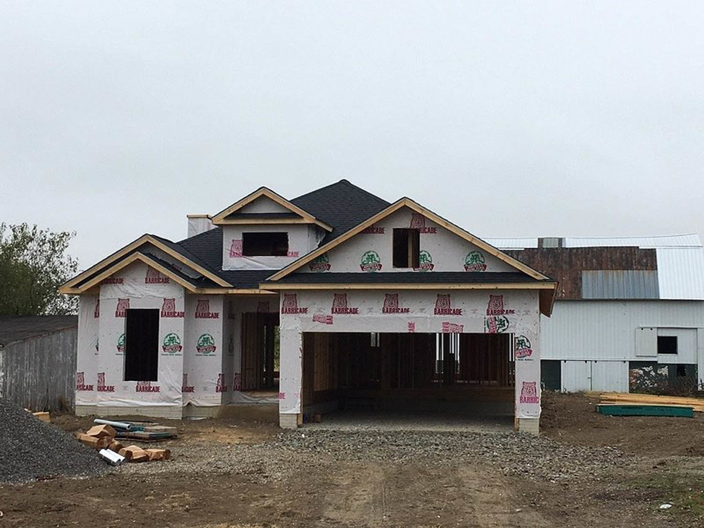 Framed house under construction with exposed plywood sheathing, attached garage, pile of metal pipes in foreground, tree beside roofline, clear sky overhead