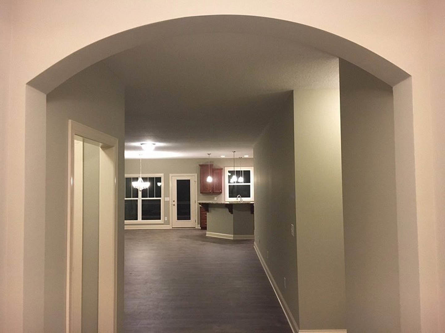 Hallway with dark wood flooring and white walls, leading to a kitchen and dining area; white door with black trim, window emitting natural light, recessed ceiling fixtures