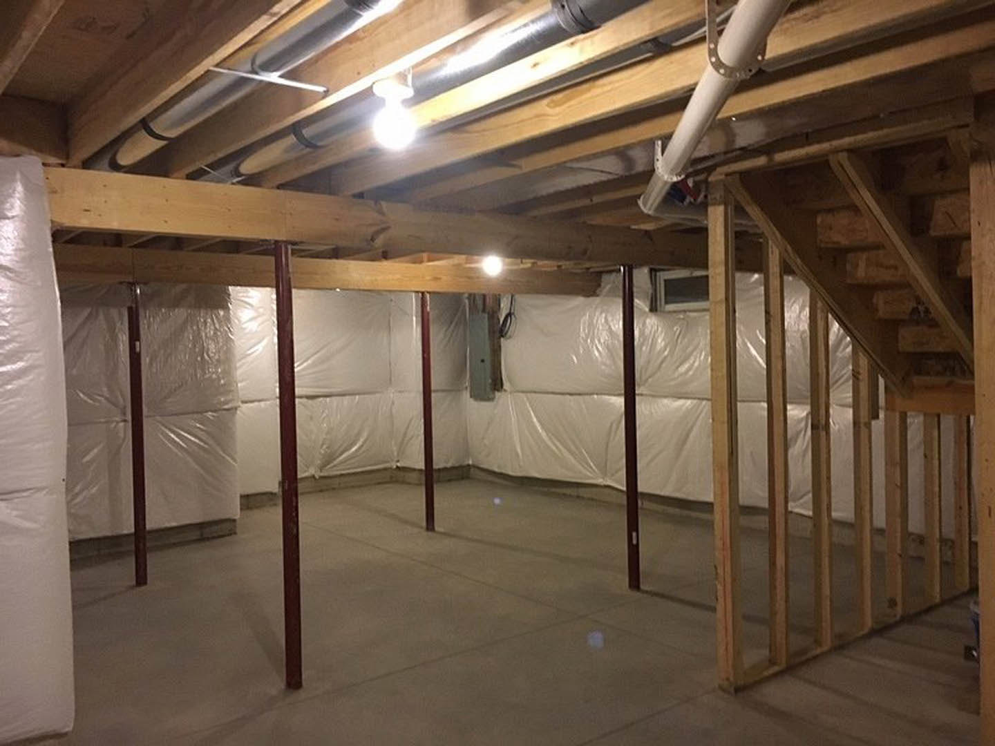 Basement with exposed wood beams, concrete floor, and white plastic sheeting covering walls and staircase; overhead light illuminates plumbing pipe and roll of toilet paper.