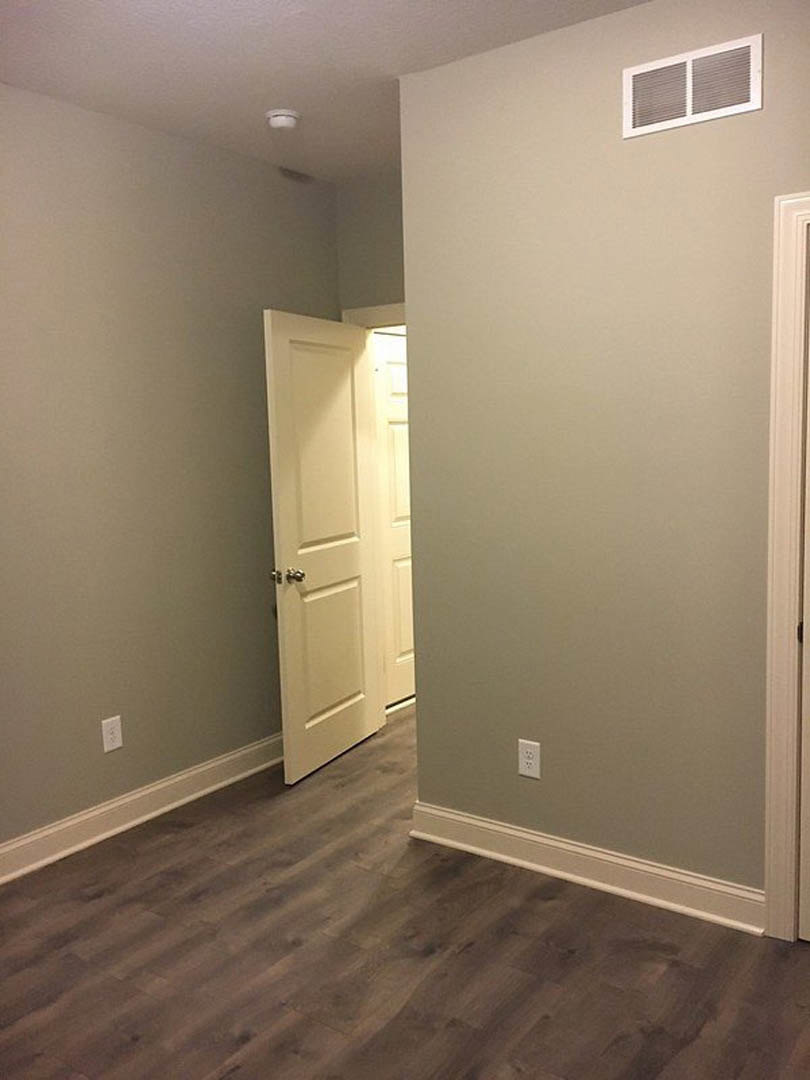 White paneled door with chrome handle set in a white wall, wood laminate flooring and white baseboard trim, rectangular wall vent, minimalist interior detailing