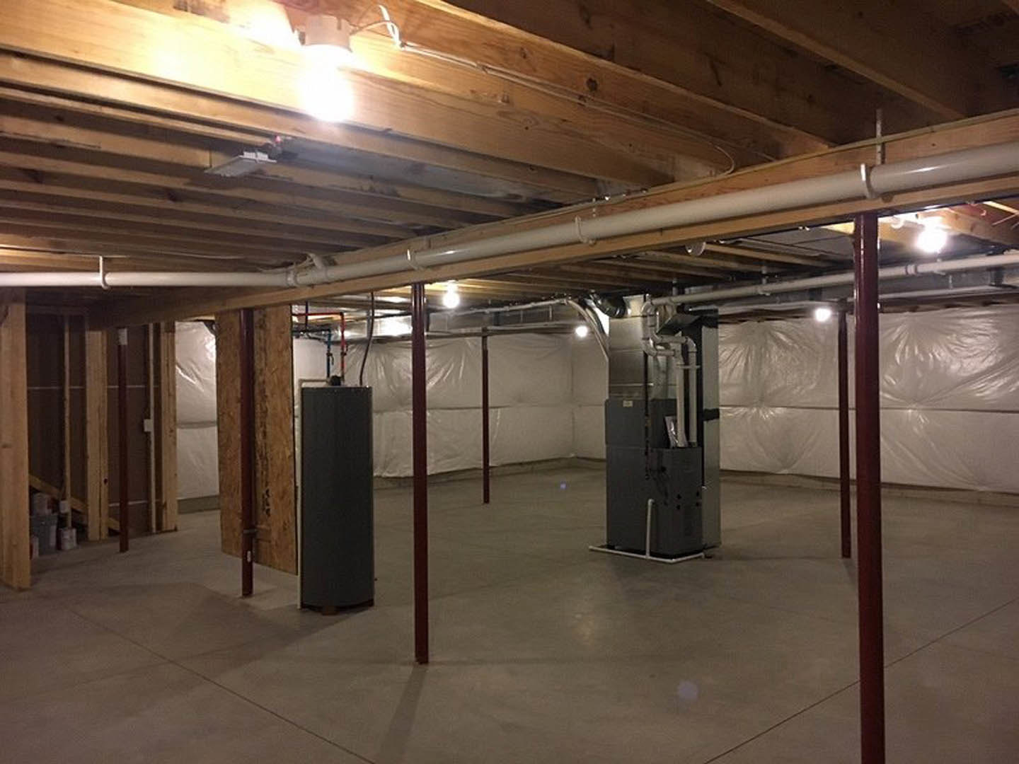 Concrete-floored utility room with exposed pipes, metal heater unit, white wall panels, and black rectangular appliance