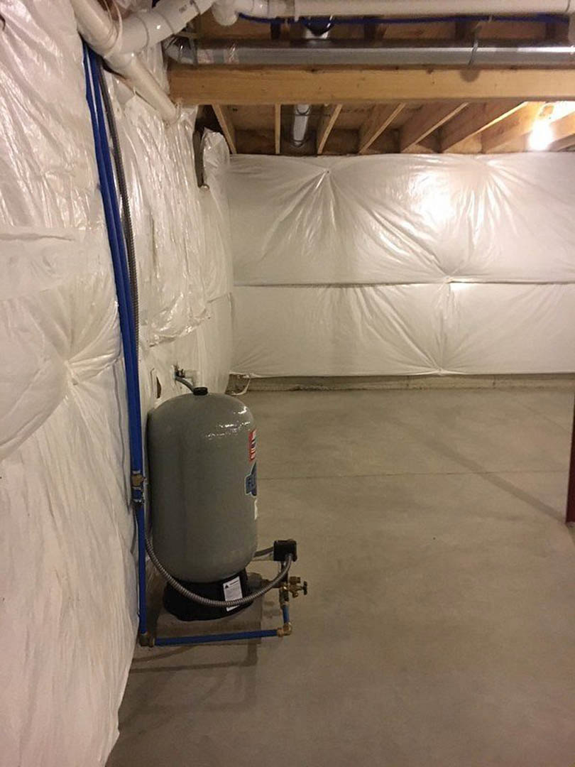 Large grey cylindrical tank with attached pipes and hose on concrete basement floor, exposed ceiling pipes, white mattress with cover nearby.