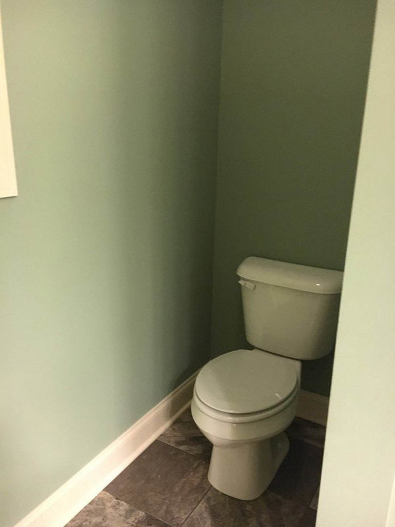 White porcelain toilet with closed lid, set against light gray tiled wall and floor, chrome flush handle, adjacent bidet and towel rack visible.
