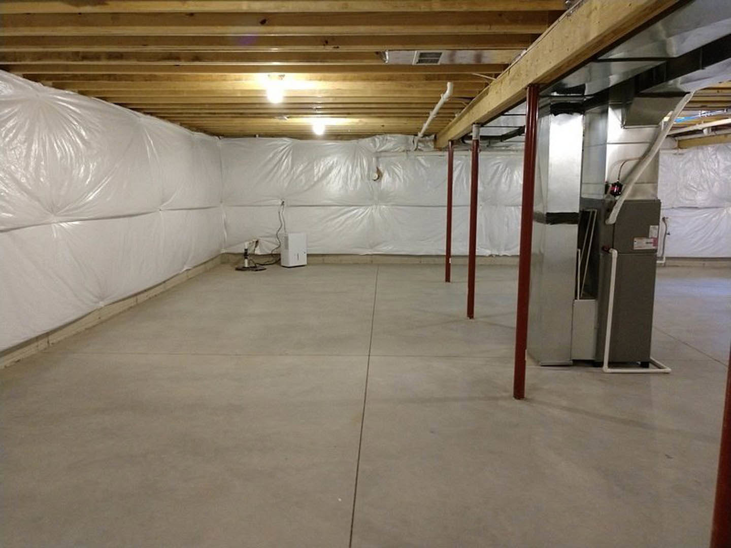 Concrete basement room with white plaster walls, metal machine with black handle, ceiling light, and white floor cover.