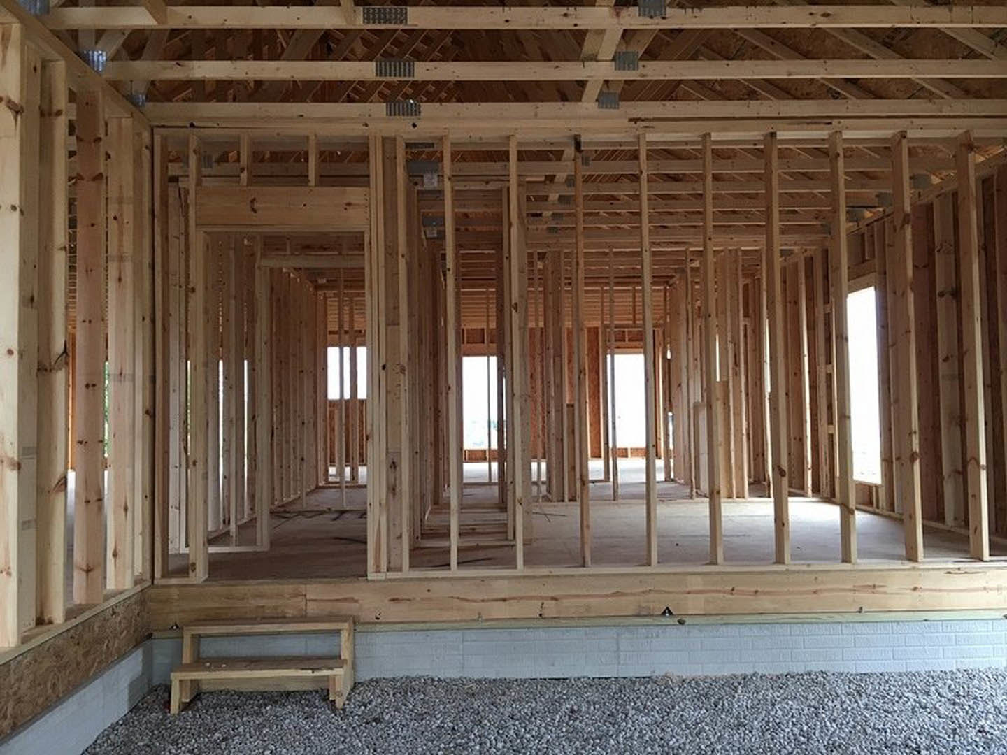 Partially finished home interior with exposed wooden beams, white brick wall, unfinished windows, stacked lumber, and gravel pile on concrete floor
