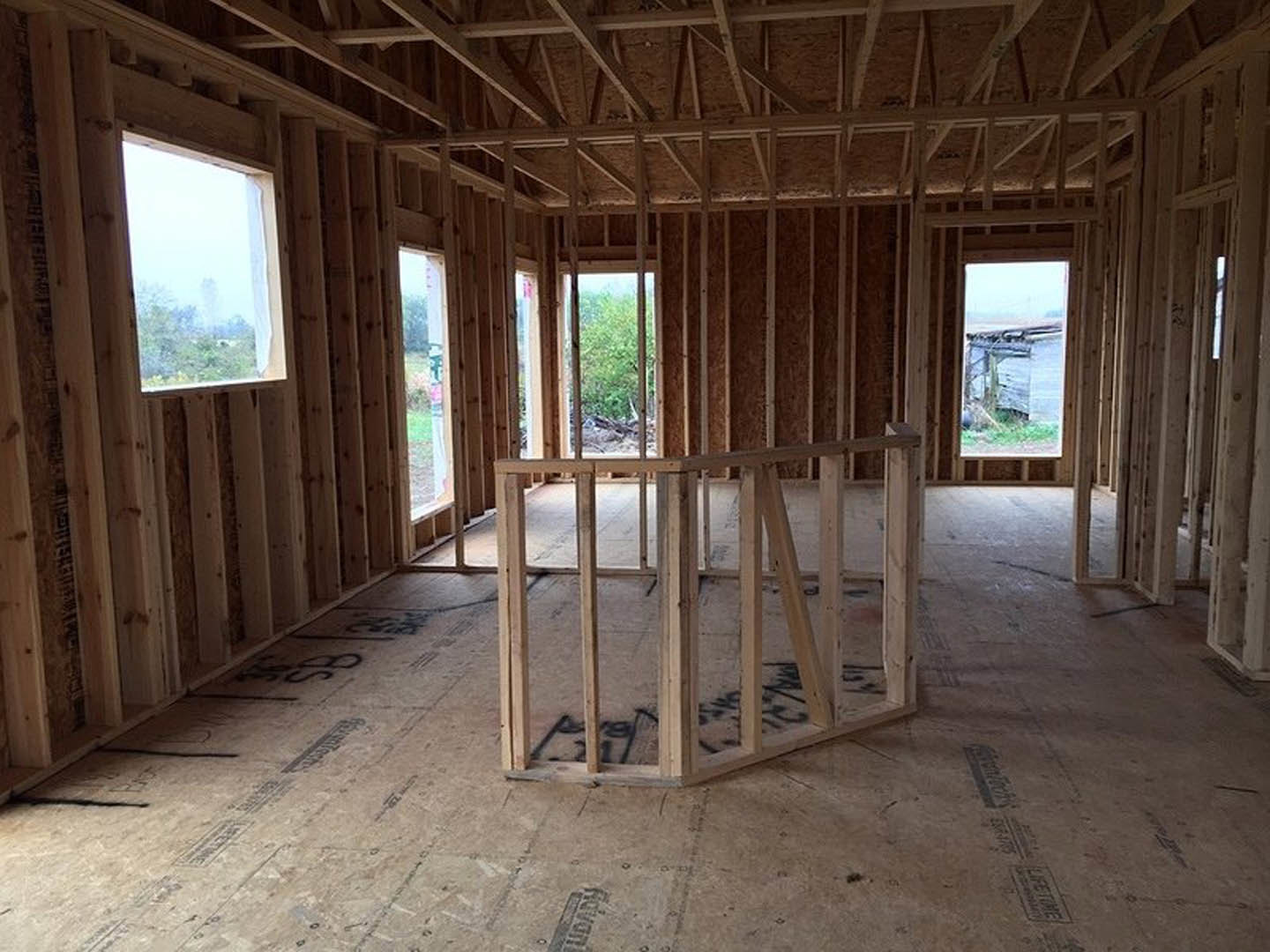 Wood-framed room under construction with exposed beams, unfinished plank flooring, and large window overlooking green trees and bushes