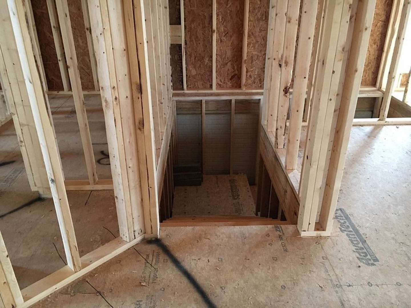 Unfinished room with exposed wood framing, plywood walls, staircase, and a hole in the wall; brown cardboard box on floor