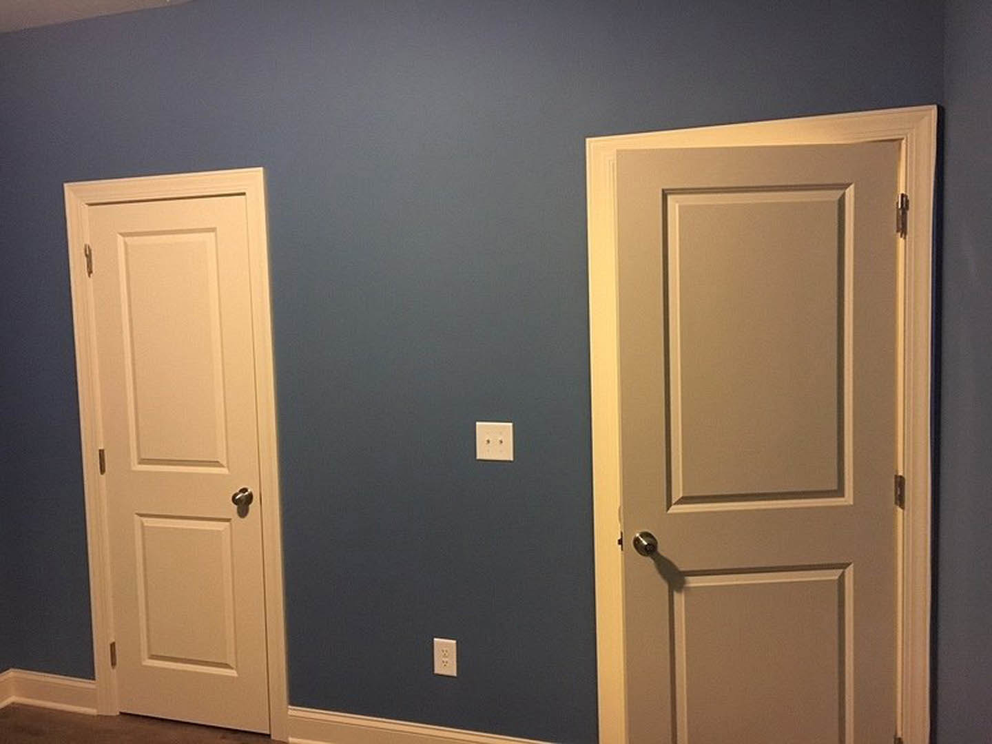 White-painted room with two matching doors featuring silver hardware, white electrical outlet on wall, light-colored flooring