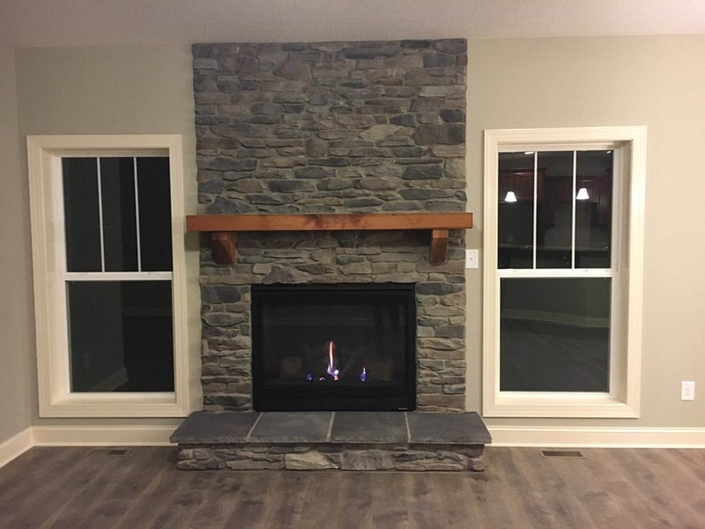Stone fireplace with wood mantel, burning fire, white-framed window with lights, hardwood floor, and stone wall in a cozy den.
