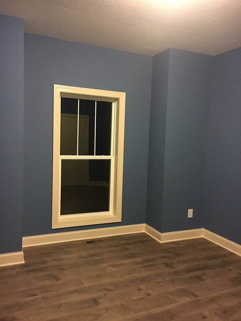 Wood floor with rectangular vent, white walls with black trim, blue accent wall, white-framed window, white door with matching handle, white ceiling.