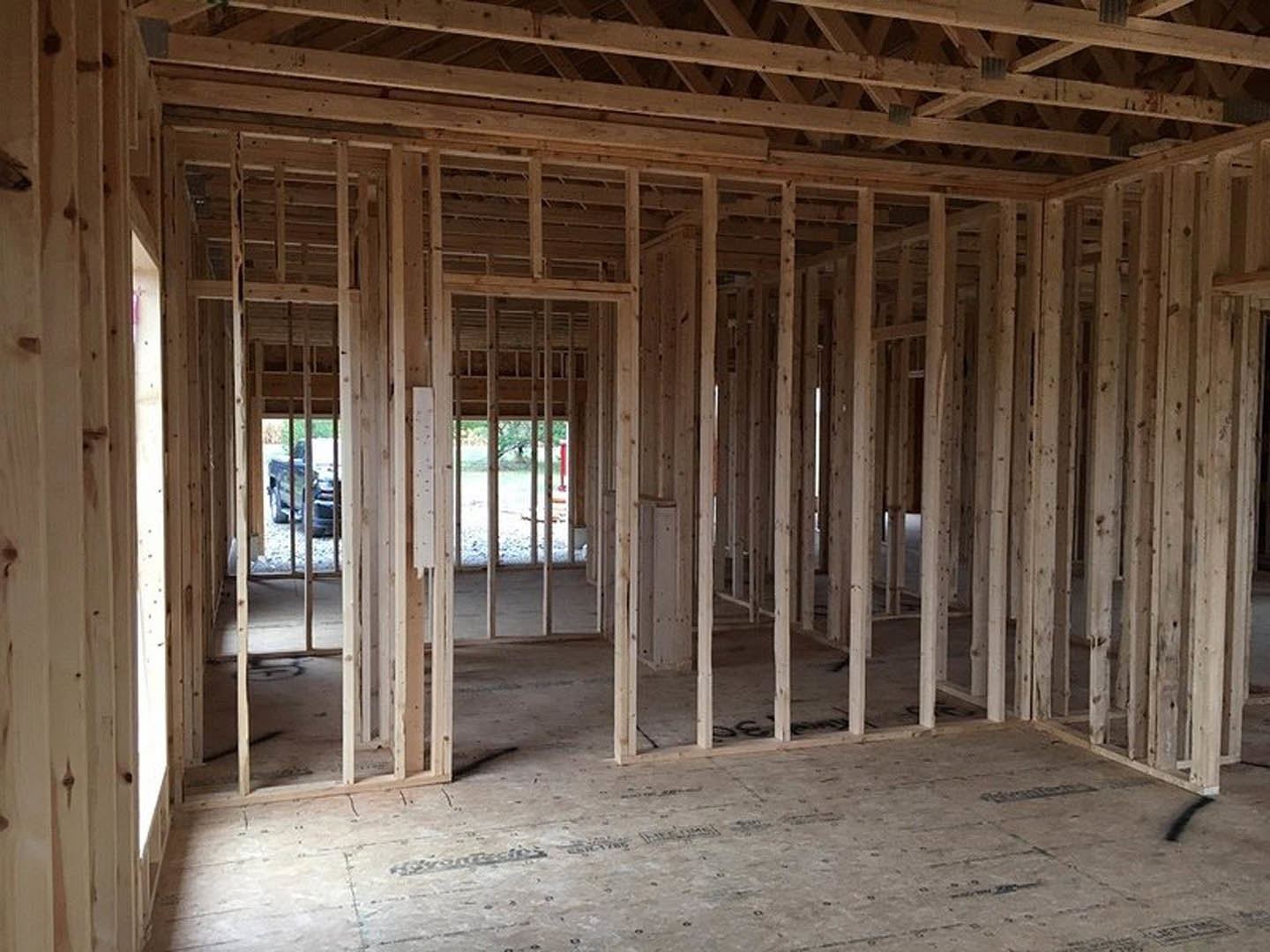 Wood-framed room under construction with exposed ceiling beams, unfinished floor, close-up of door, black line marking on ground, and window frame with metal bars
