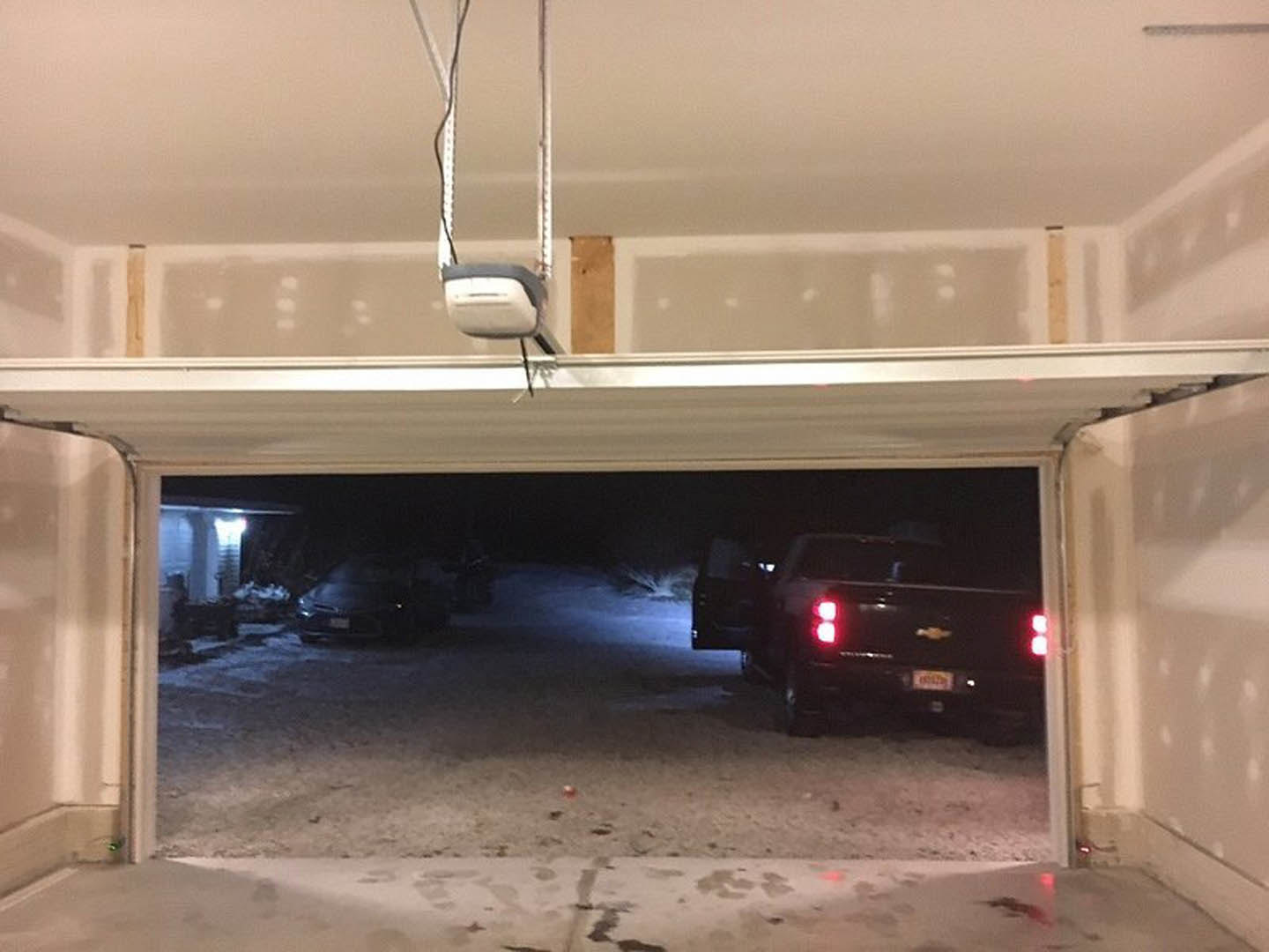 Garage interior with white walls, closed garage door, black truck with red tail lights parked inside, white metal shelving, concrete floor, and visible ceiling lighting.