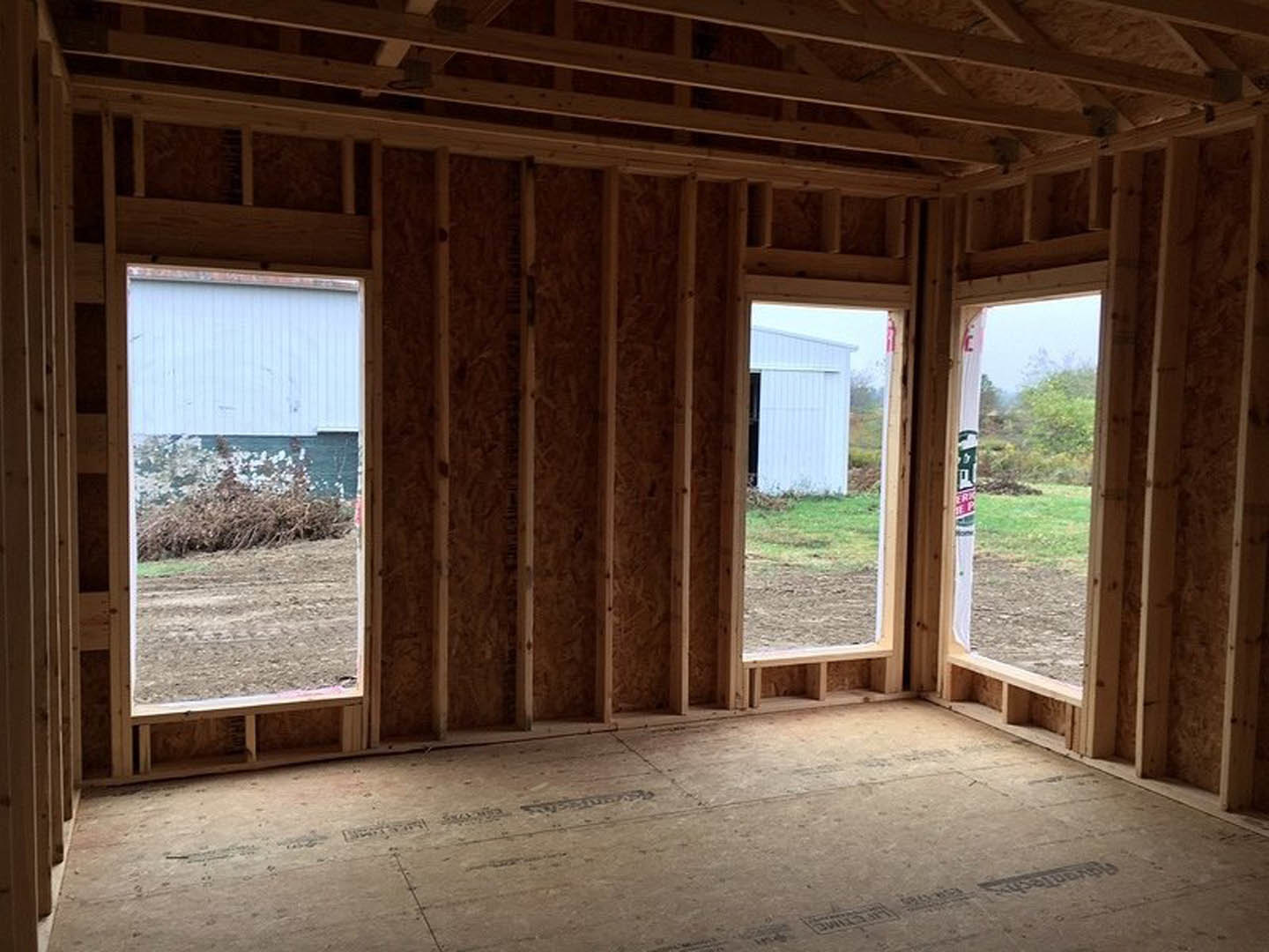 Room under construction with exposed wood framing, multiple windows, unfinished floor, and view of green field and trees through the glass.