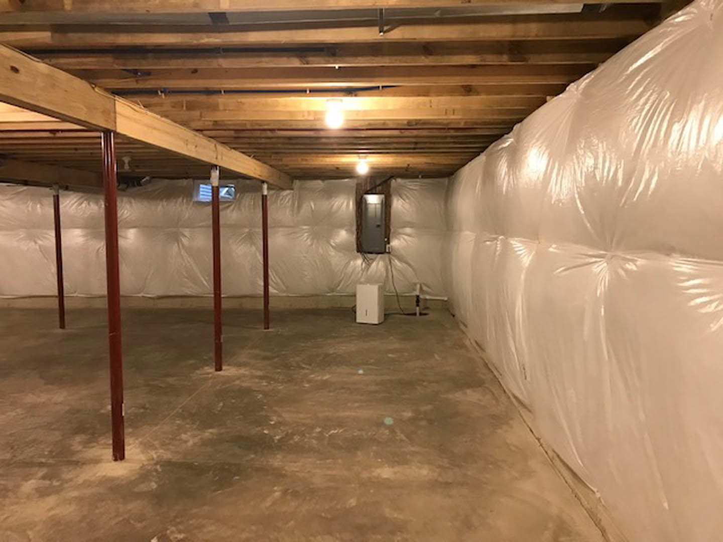 Basement with white plastic wall covering, exposed wooden ceiling beams, large white bag on floor, light bulb hanging from ceiling, white rectangular object with black handle near