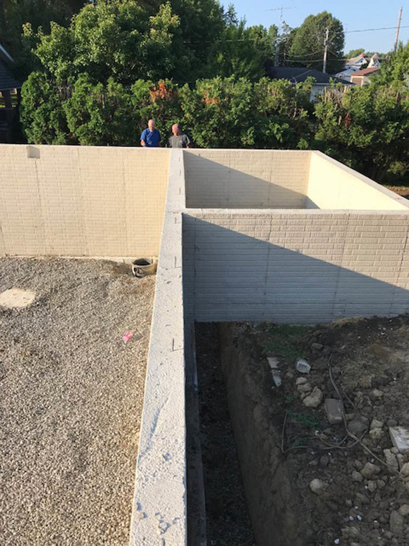 Two men standing on a concrete wall near gravel and dirt ground, with trees and plants in the background under a partly cloudy sky