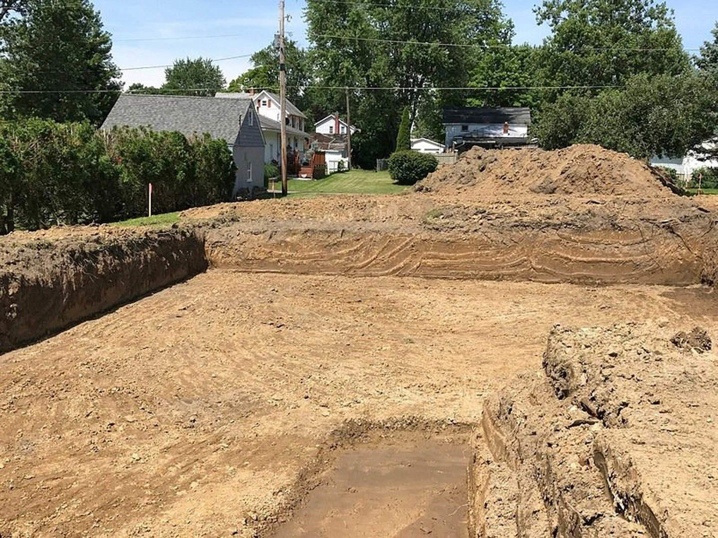 Excavated dirt pit with exposed soil and rocks, surrounded by green bushes and trees, partially visible house in background under open sky