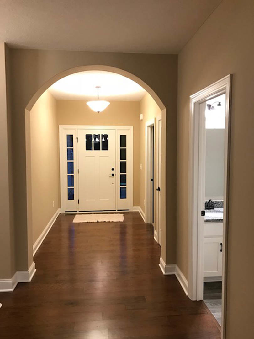 Hallway with hardwood floor, white door featuring glass panes, patterned rug, ceiling light fixture, view through doorway into kitchen