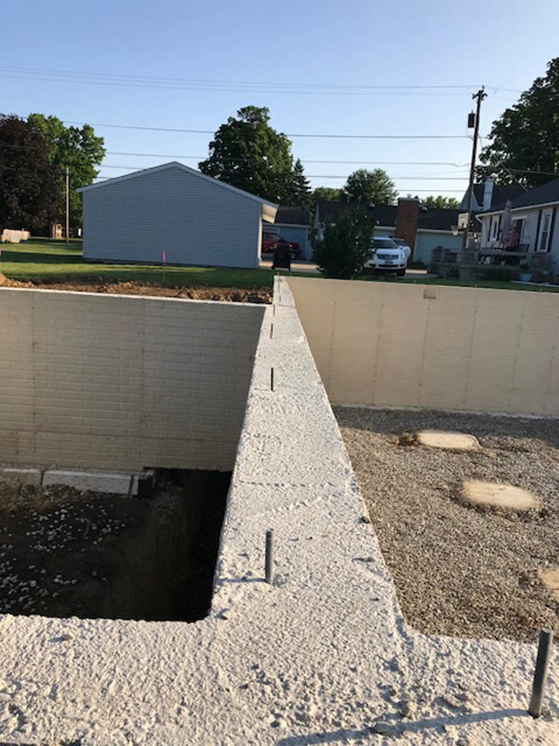 Concrete exterior wall with circular hole in the ground, grey house with pink pole, tree with green leaves, dirt ground, white car in background, person standing near white wall