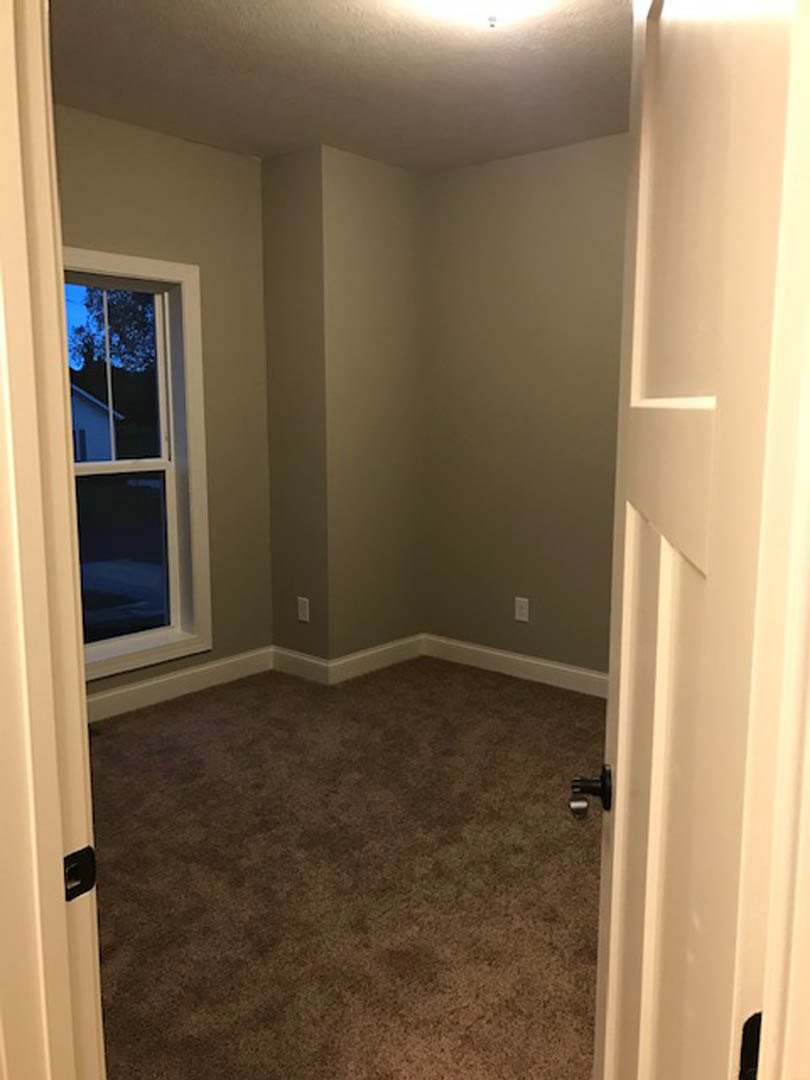 Brown carpeted floor in a room with a white door open, window reflecting a neighboring house, black wall fixture, and plaster walls.