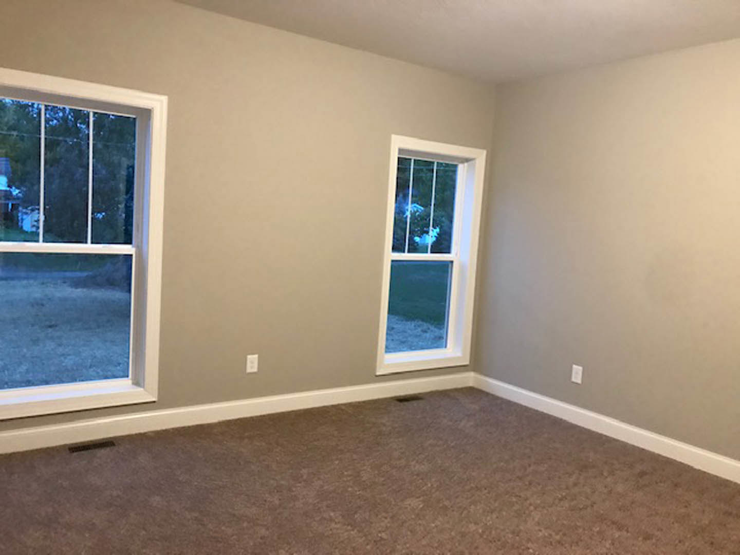Spacious carpeted room with two large white-framed windows, neutral walls, and natural light illuminating the interior