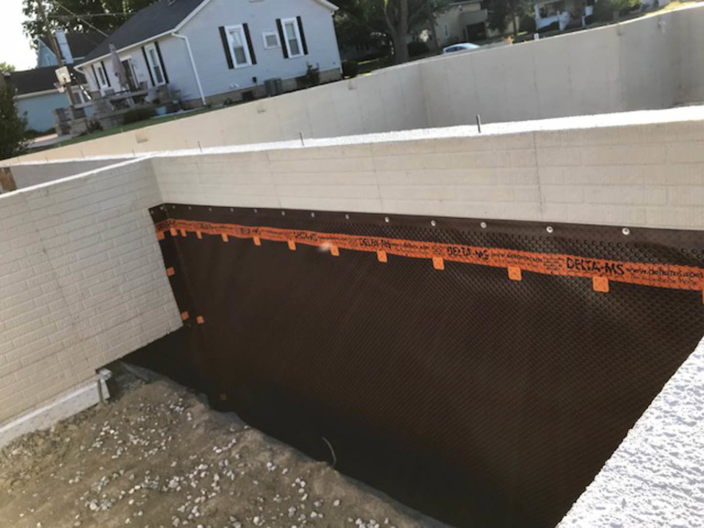 Concrete house foundation with brown metal formwork, orange tape marking edges, surrounded by rocky ground and trees under a cloudy sky.