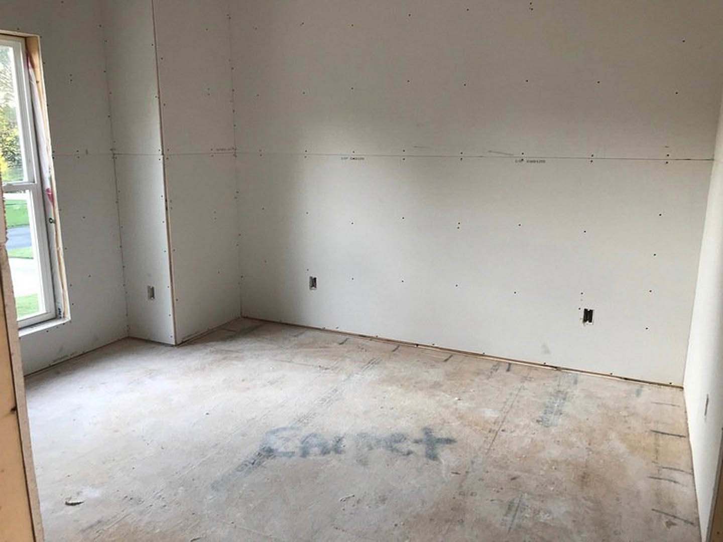 Unfinished room with white plaster walls featuring multiple holes, concrete floor marked with writing, and a window with a white frame