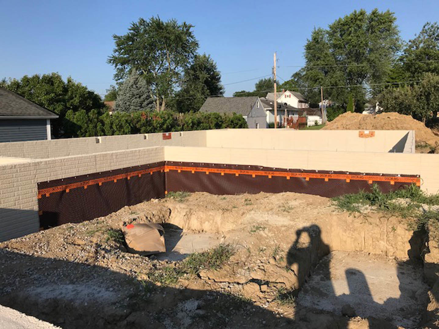 Concrete foundation excavation surrounded by dirt and construction barriers, partially built house with garage door in background, blue sky and trees visible