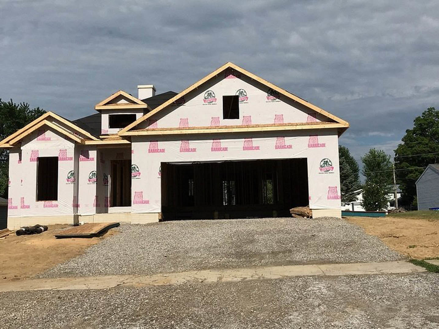 Two-story house under construction with exposed plywood sheathing, pink inspection stickers, white-framed windows, and a gravel driveway in front