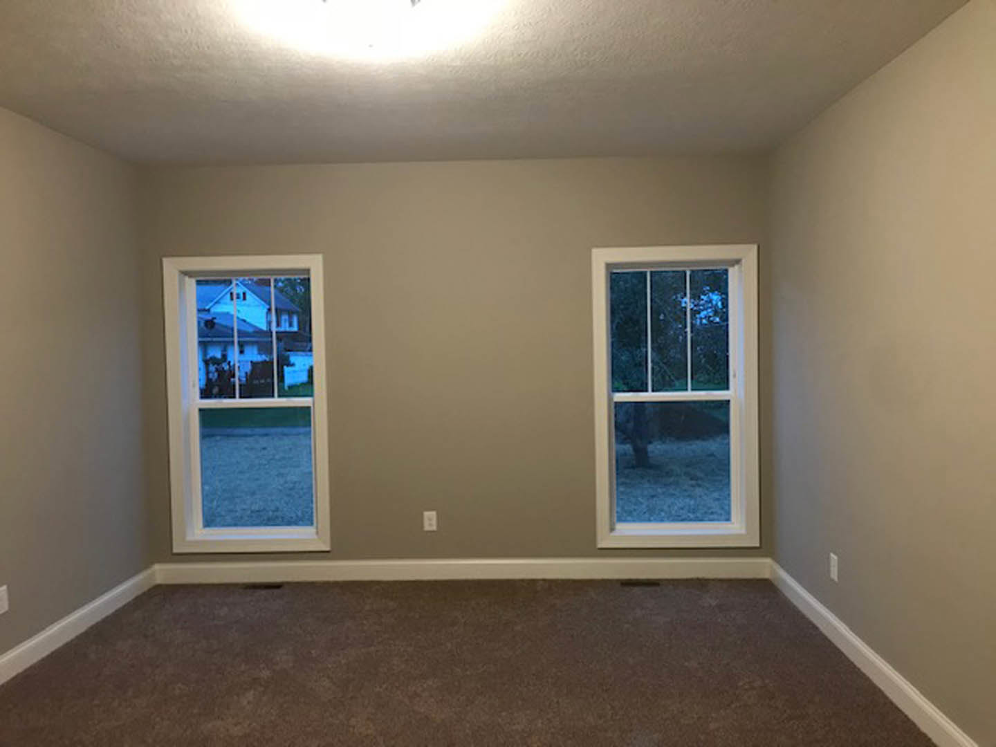 Bright room with two large windows, neutral plaster walls, carpeted floor, and views of neighboring houses and sky.