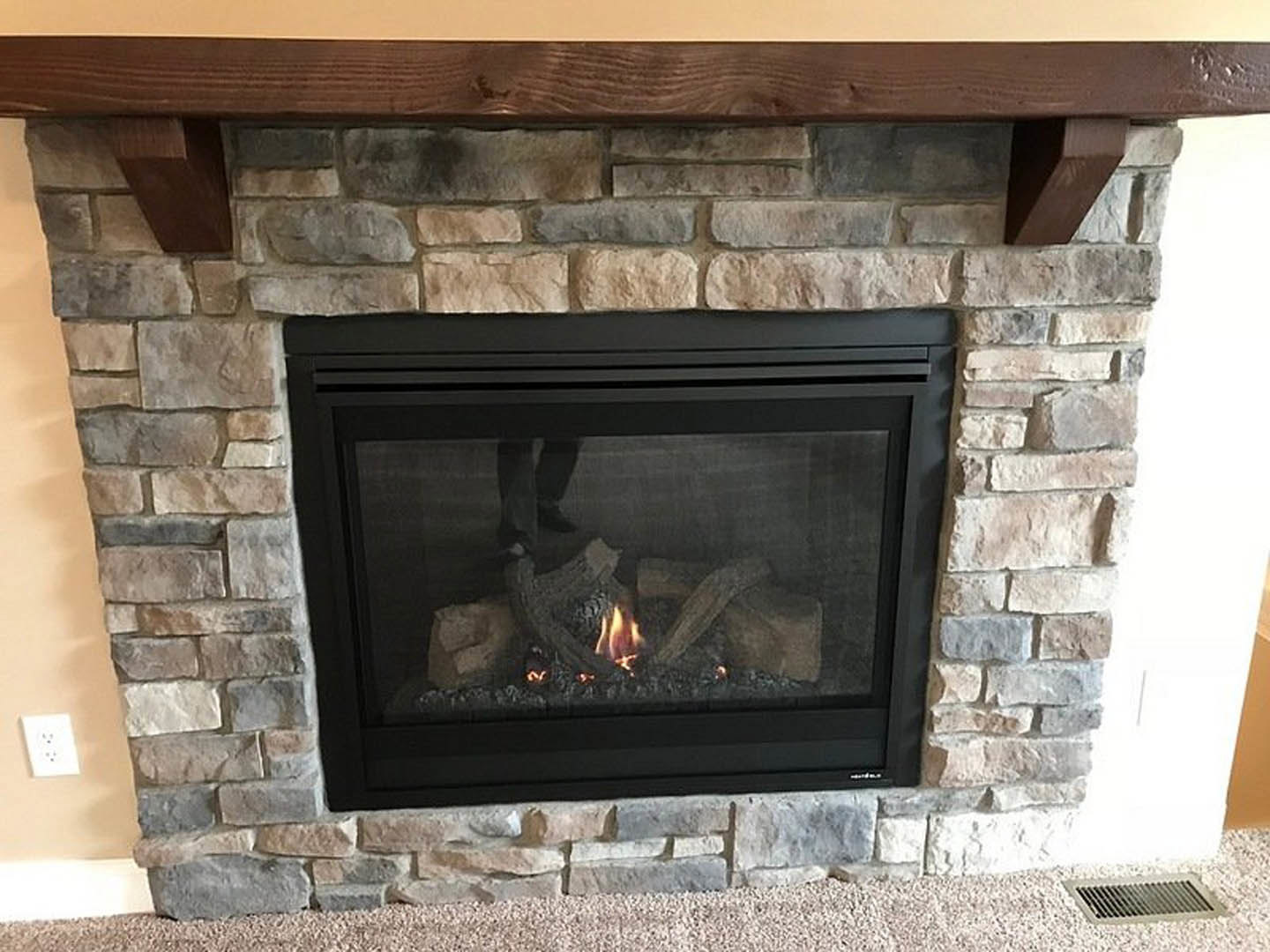 Stone fireplace with wood burning inside, brick hearth, fire screen, and partial view of a person's leg; close-up of masonry wall and white electrical outlet with black dots