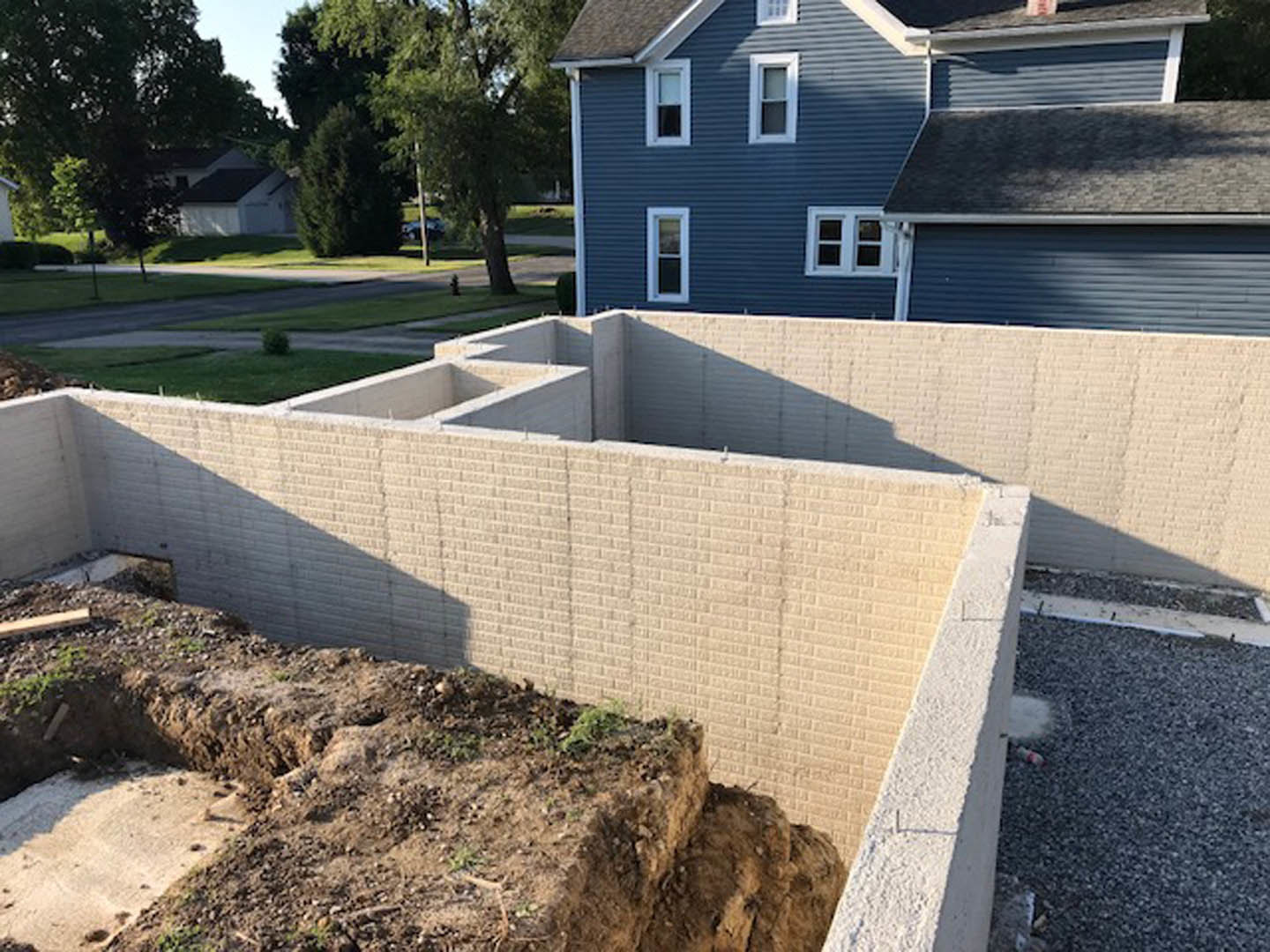 White brick house with a white roof, surrounded by trees, adjacent to a dirt patch and construction site with exposed concrete and a window with a white frame.