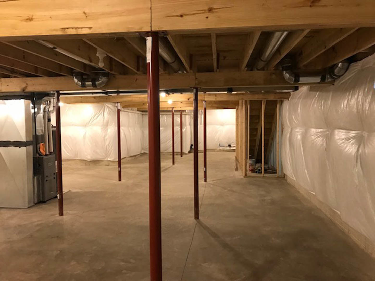 Basement with exposed wooden ceiling, visible pipes, concrete floor with red support poles, white bag attached to wall, metal and wooden beams, close-up of metal pole