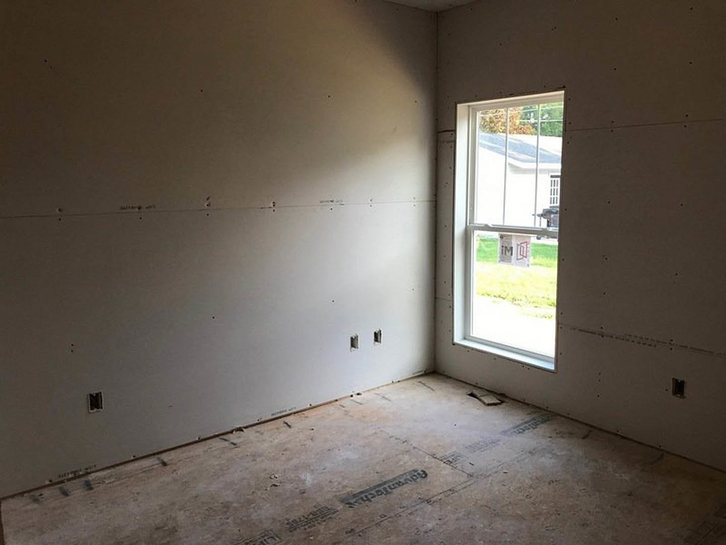 Concrete floor with visible seams, plaster walls, large window framed in wood, minimal ceiling fixtures, natural light illuminating sparse interior
