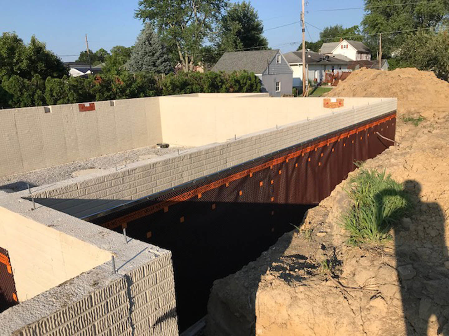 Construction site with exposed trench bordered by temporary fencing, stone retaining wall, patches of grass, and concrete foundation elements visible.