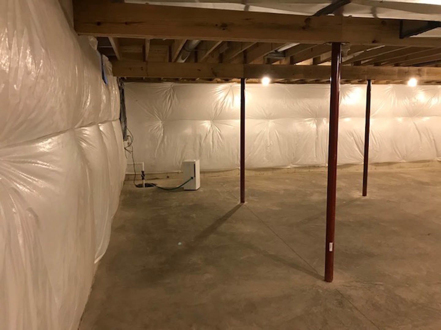 Basement with white plastic wall covering, exposed wooden ceiling beams, concrete floor, white storage boxes, and a bed with a white pillow.