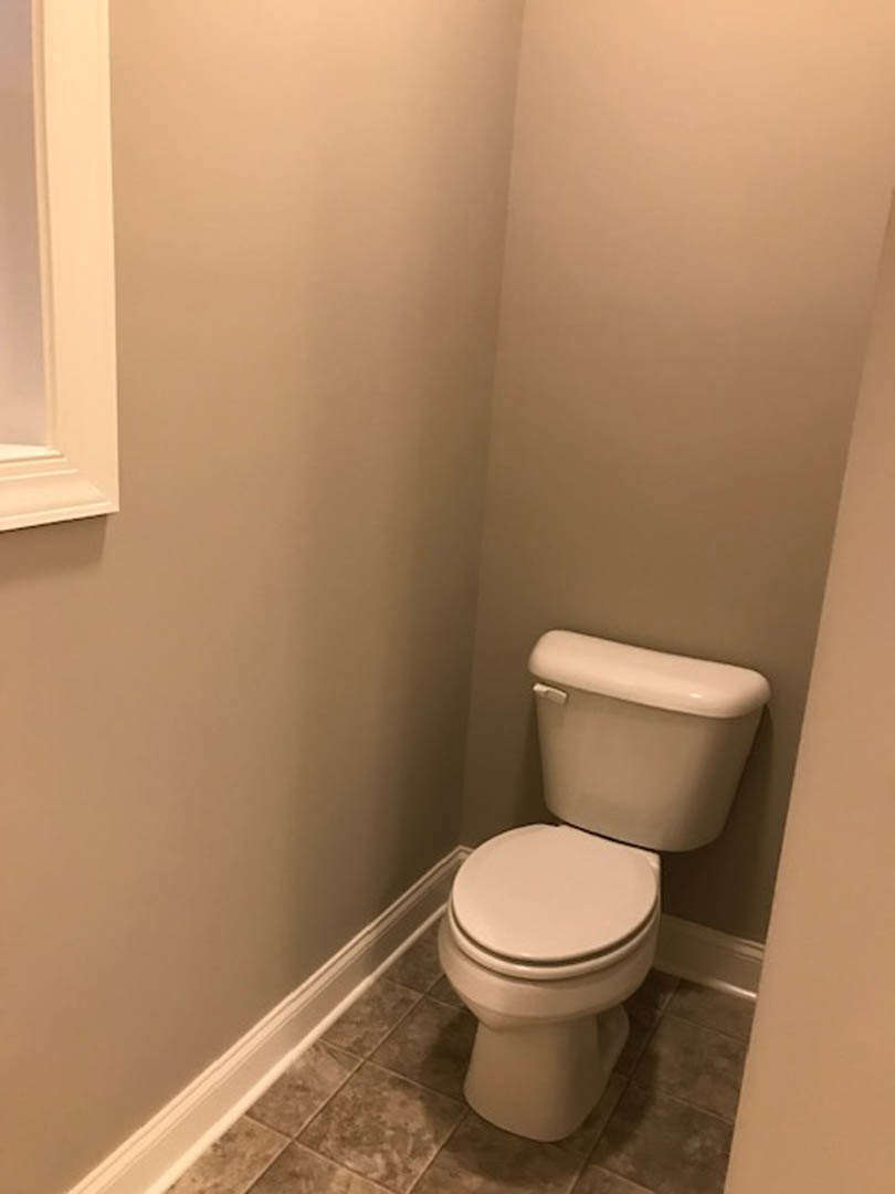 White porcelain toilet with chrome handle set against a smooth white wall, gray tile flooring visible beneath, soft shadow cast along the base.
