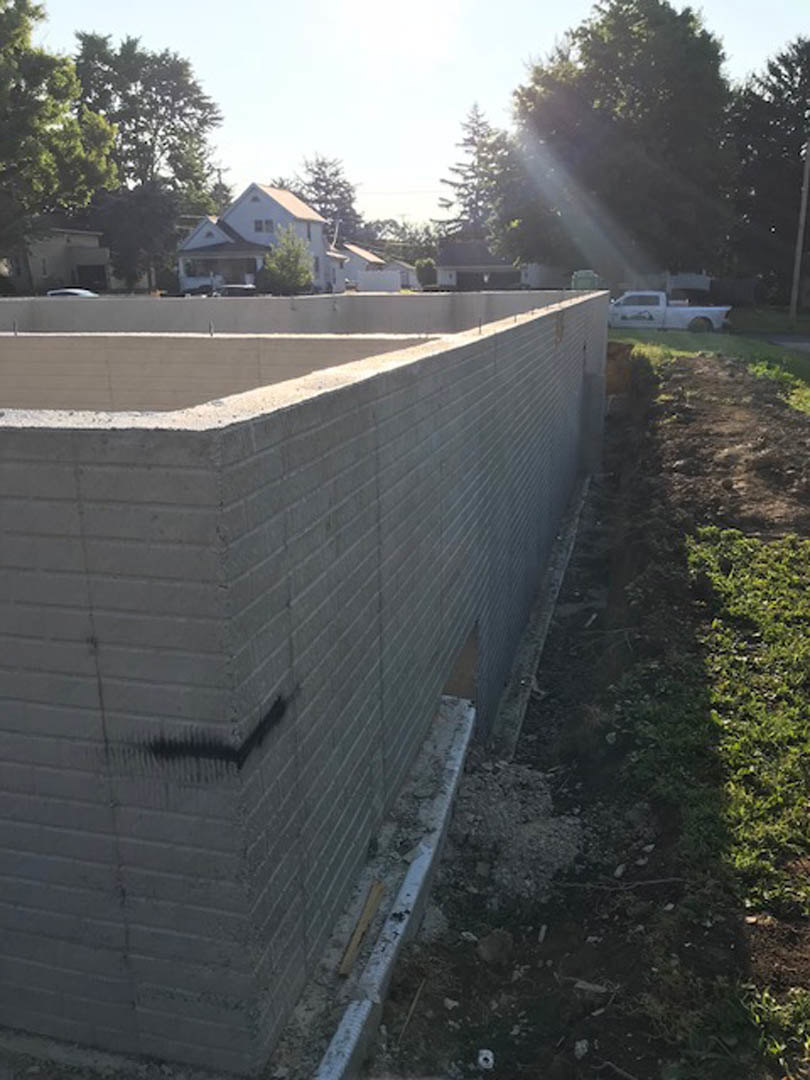 Partially built brick house with exposed concrete walls, construction materials scattered in front, several parked cars, and mature trees surrounding the site.