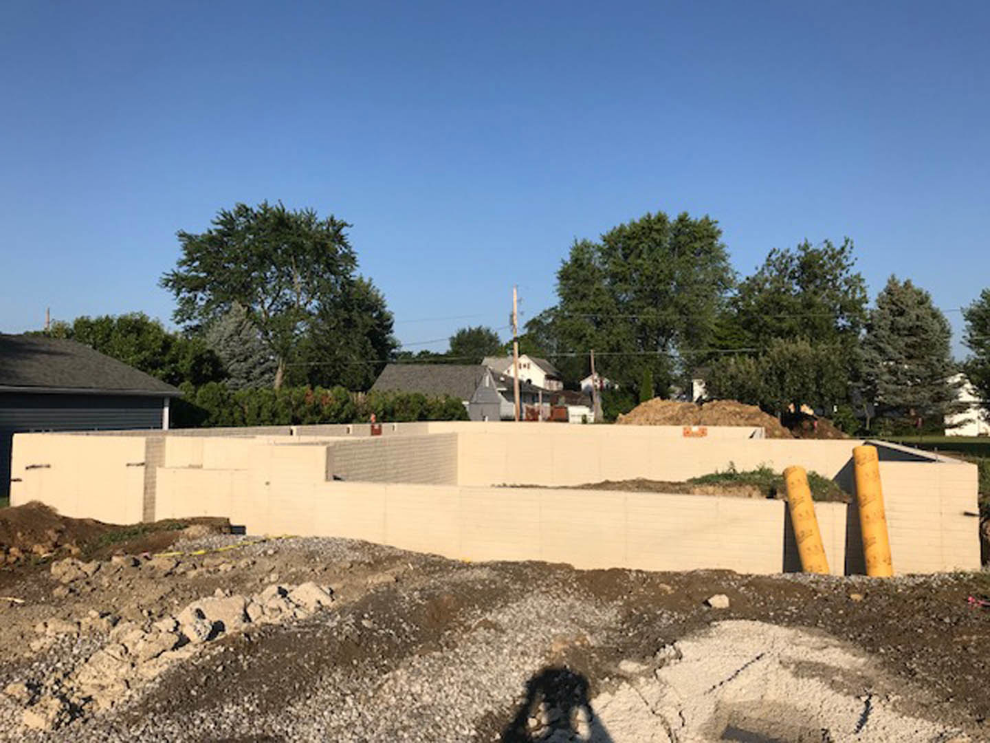 Framed custom home under construction with exposed white walls, central opening, dirt ground, nearby trees, and neighboring houses under blue sky